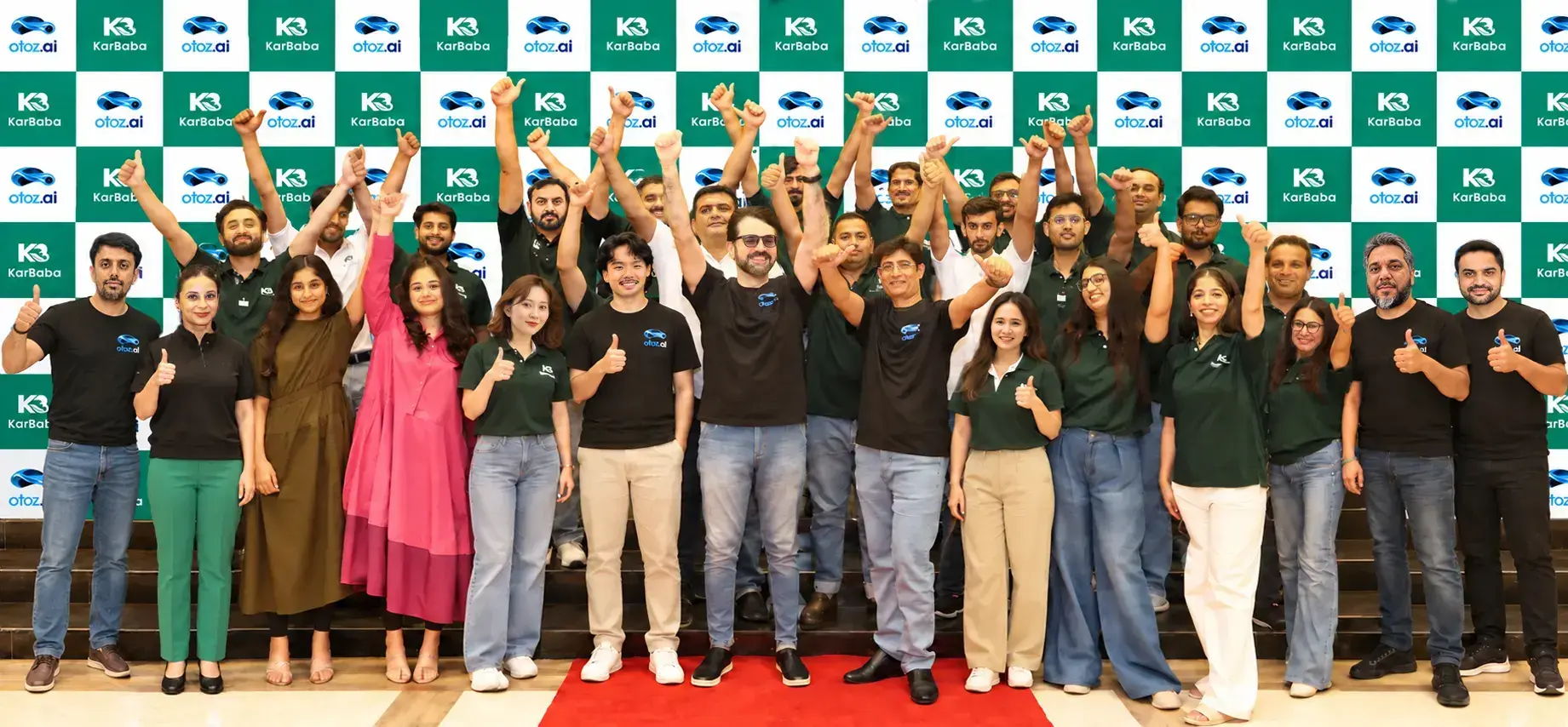 The KarBaba and otoz.ai team cheering with thumbs up in front of a branded backdrop